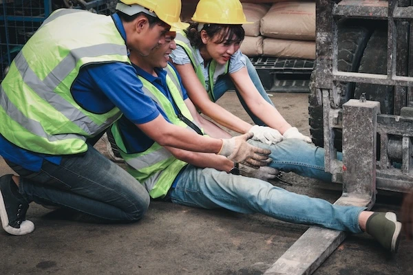 two factory workers trying to help another worker with his leg stuck in a forklift